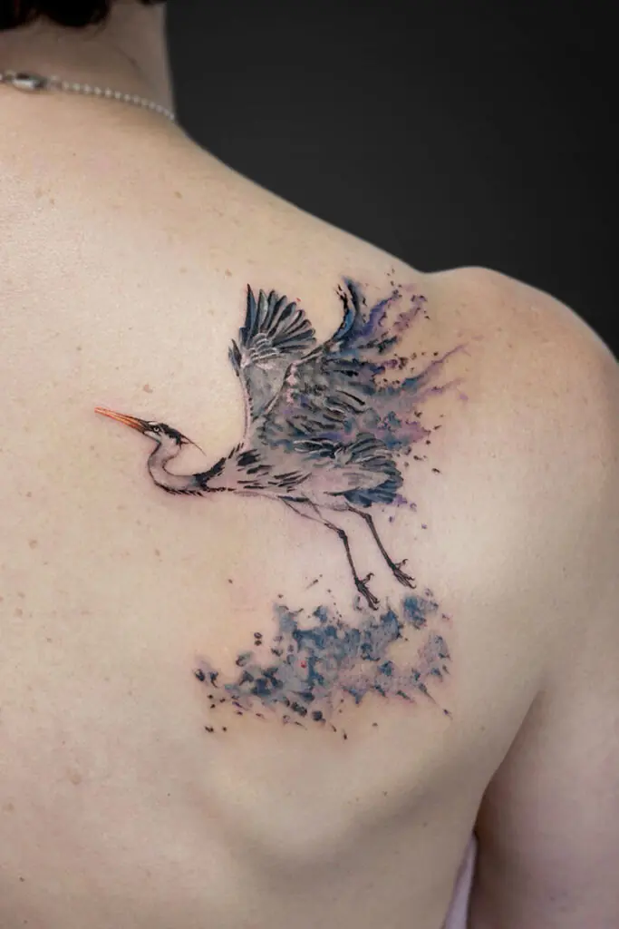 Watercolor-style tattoo of a heron in flight on a person’s upper back shoulder, with blue-purple splashes behind.