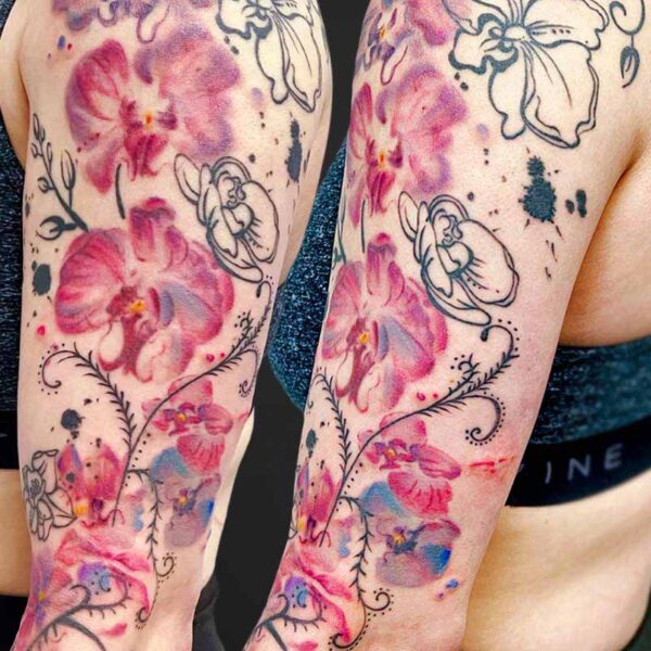Watercolor floral tattoo sleeves on both arms, with pink, purple, and blue blooms and black vine outlines extending down the forearms.