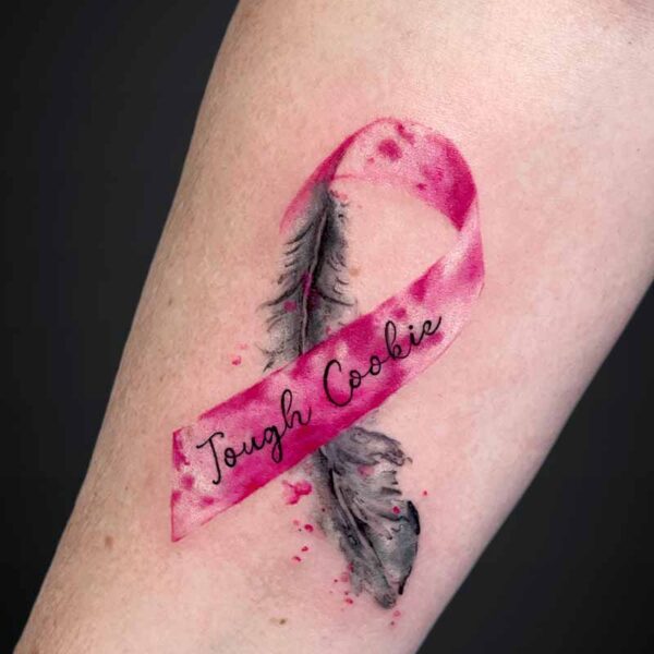Watercolor pink ribbon tattoo on forearm with a black feather and the cursive text 'Tough Cookie'.