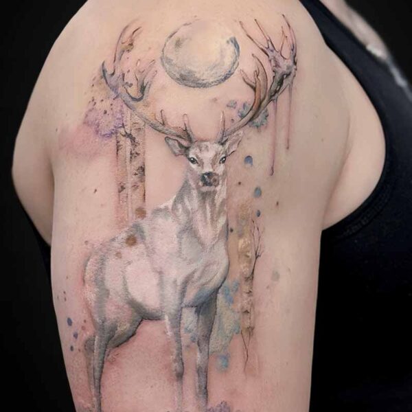 Watercolor-style deer tattoo on the upper arm: a white stag with large antlers beneath a pale moon, with soft pastel splashes around.