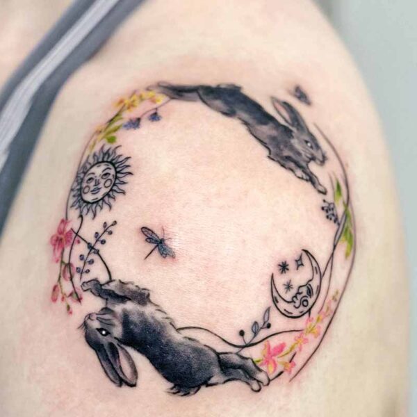 Circular tattoo on upper arm featuring two black rabbits, sun and moon motifs, and scattered flowers in a delicate watercolor style