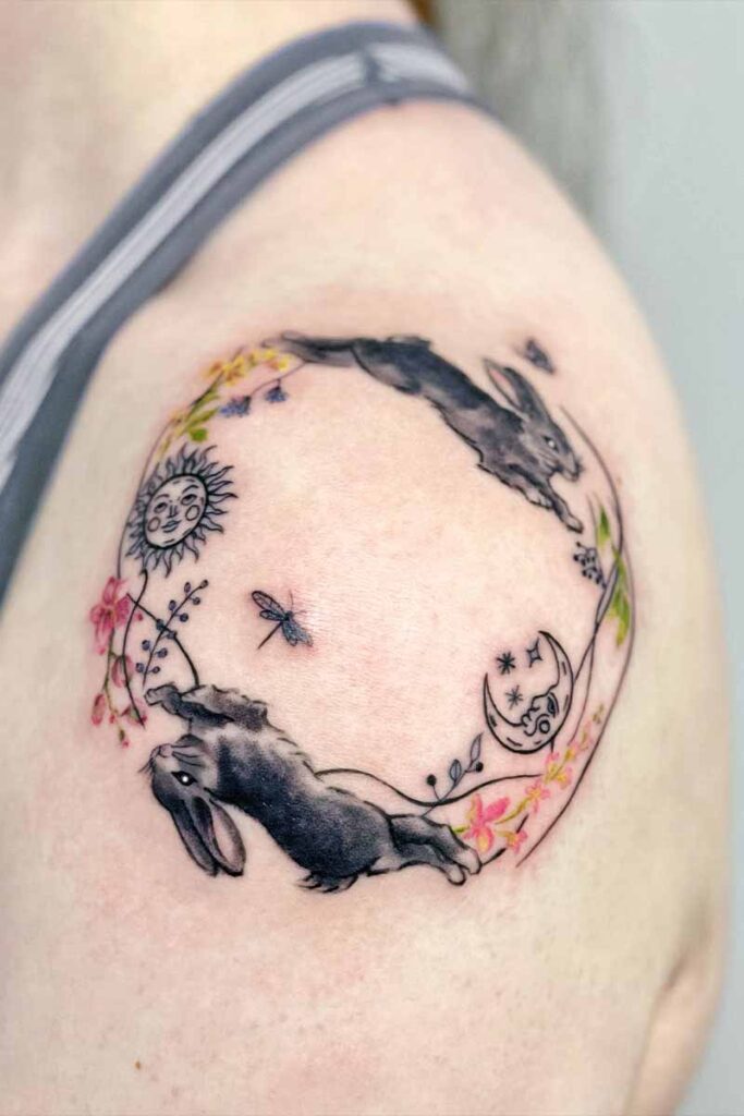 Circular tattoo on upper arm featuring two black rabbits, sun and moon motifs, and scattered flowers in a delicate watercolor style